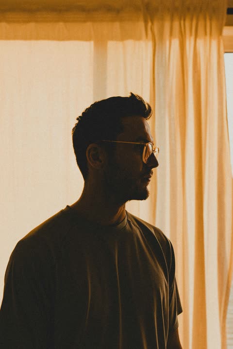 A bearded man in profile beside a bright curtain