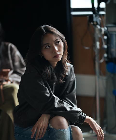 A woman seated in a dim studio, looking toward soft window light